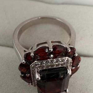 May include: A silver ring with a large, rectangular, dark red gemstone at its centre, encircled by smaller, oval red gemstones. The ring has a detailed setting with small, clear accent stones.