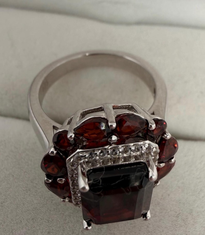 May include: A silver ring with a large, rectangular, dark red gemstone at its centre, encircled by smaller, oval red gemstones. The ring has a detailed setting with small, clear accent stones.