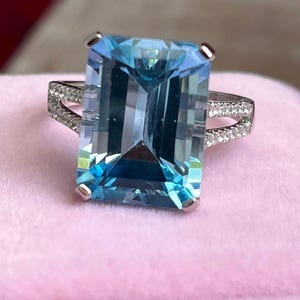 Puede incluir: Anillo de piedra preciosa rectangular azul claro con una banda plateada. La piedra preciosa es una piedra grande, clara y facetada. La banda presenta pequeñas piedras de acento claras. El anillo está sobre una superficie rosa.