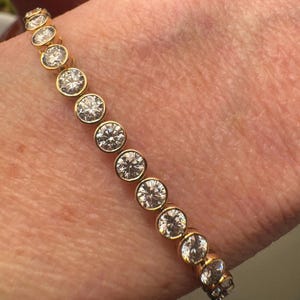 A gold bracelet with round, clear Moissanite stones set in a bezel setting.