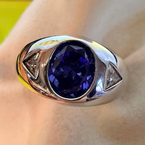 May include: A silver ring featuring a large, oval-cut deep purple gemstone set in the center, flanked by two smaller, triangular-cut clear stones on either side. The band is smooth and polished, with a wide, rounded profile surrounding the stones.