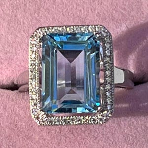 May include: A rectangular aquamarine gemstone ring with a halo of small, clear stones. The aquamarine is a light blue color with a rectangular cut. The ring is set in a silver band.