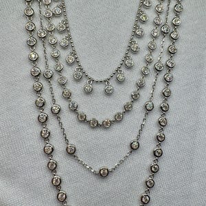 May include: A silver multi-strand necklace with round, clear stones. The necklace has a graduated length with the longest strand featuring a dangling design.