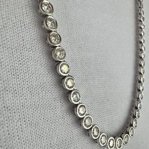 May include: A silver necklace with a series of round, clear gemstones set in individual bezels. The gemstones are evenly spaced and create a continuous line. The necklace is displayed against a white, textured background.