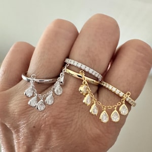 May include: A hand displays three rings: a silver ring with dangling teardrop-shaped gems, and two gold rings, one with small gems and the other with dangling teardrop gems. The rings are set against a neutral background.