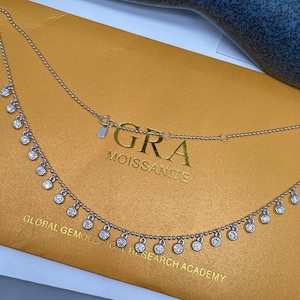 May include: A silver necklace with a delicate chain and small round stones hanging from it. The necklace has a tag that reads "GRA MOISSANITE GLOBAL GEMOLOGICAL RESEARCH ACADEMY".