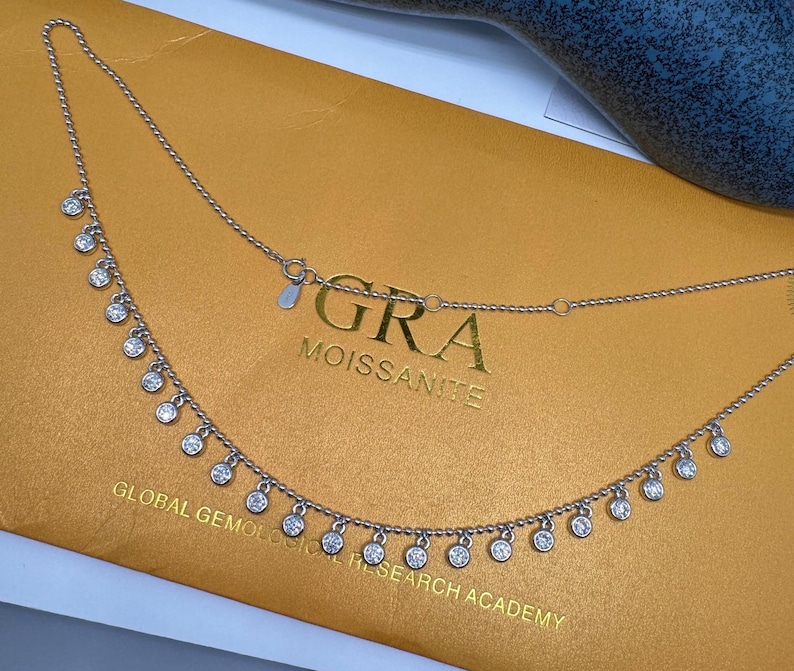 May include: A silver necklace with a delicate chain and small round stones hanging from it. The necklace has a tag that reads "GRA MOISSANITE GLOBAL GEMOLOGICAL RESEARCH ACADEMY".