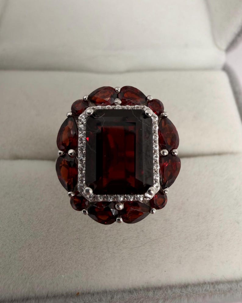May include: A silver ring with a large, rectangular, deep red gemstone at its centre, encircled by smaller, teardrop-shaped red stones. The central stone is framed by a row of small, clear stones, creating a striking contrast.