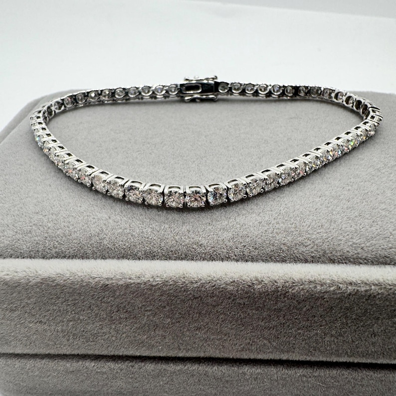 May include: A silver tennis bracelet featuring numerous round, clear gemstones. The bracelet is presented on a grey velvet surface, highlighting its brilliance. The clasp is visible, showing the bracelet's secure fastening. The bracelet is approximately 18 cm in length.