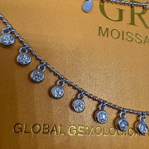 May include: A silver necklace with a delicate chain and multiple dangling round stones. The necklace is on a gold background with the text "GLOBAL GEMOLOGICAL" and "GR MOISSANITE" visible.