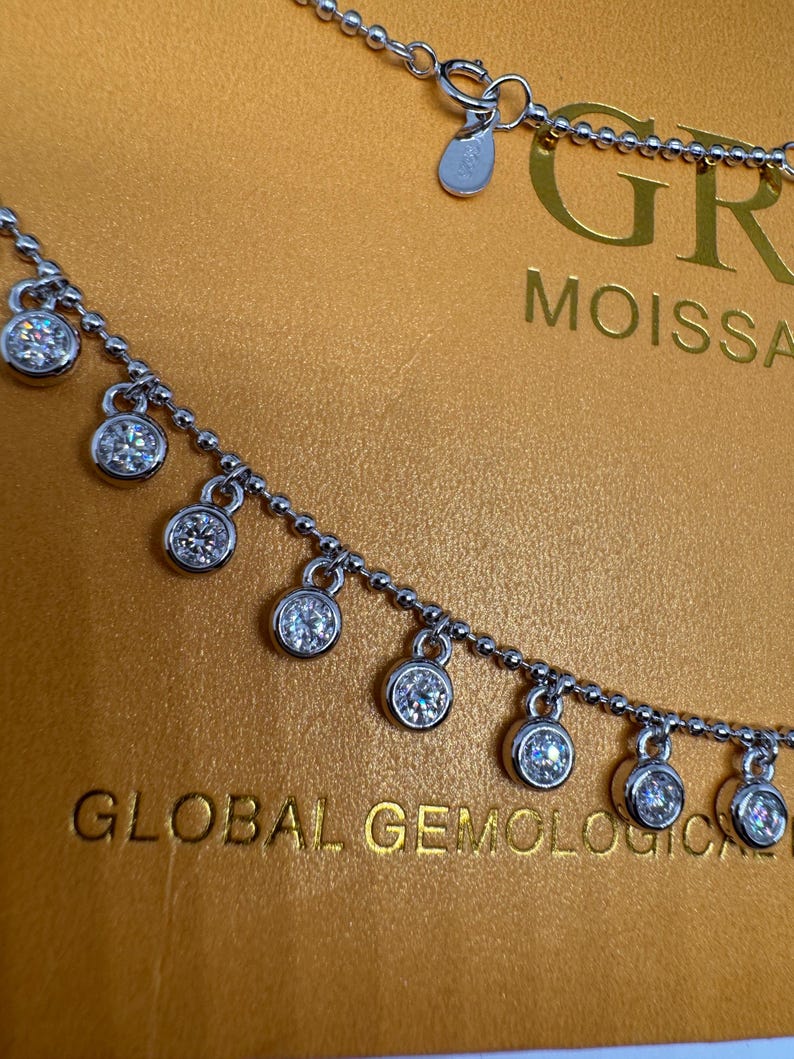 May include: A silver necklace with a delicate chain and multiple dangling round stones. The necklace is on a gold background with the text "GLOBAL GEMOLOGICAL" and "GR MOISSANITE" visible.