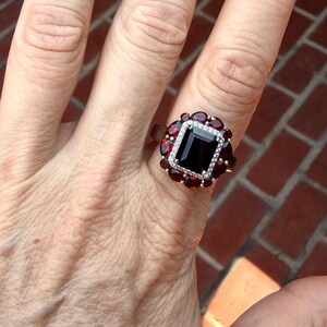 May include: A ring featuring a large, rectangular, deep red center stone surrounded by a halo of smaller, round, clear stones, which is further encircled by a cluster of pear-shaped, deep red stones set in a metallic band.