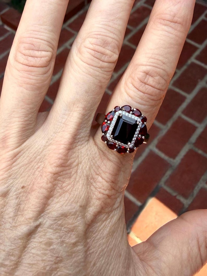 May include: A ring featuring a large, rectangular, deep red center stone surrounded by a halo of smaller, round, clear stones, which is further encircled by a cluster of pear-shaped, deep red stones set in a metallic band.