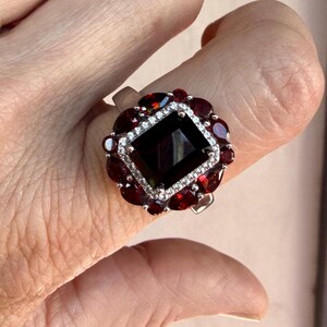 May include: A silver ring featuring a large rectangular dark red center stone surrounded by a halo of small clear stones. A secondary outer border consists of multiple pear-shaped dark red stones arranged in a floral-like pattern around the center.