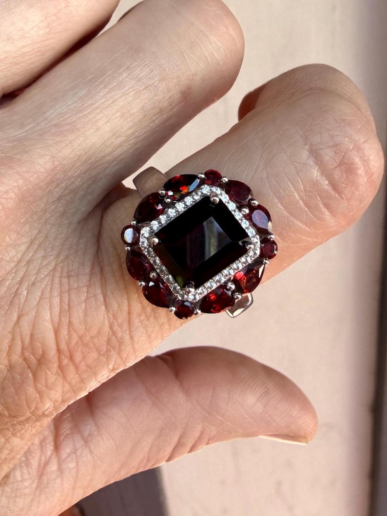 May include: A silver ring featuring a large rectangular dark red center stone surrounded by a halo of small clear stones. A secondary outer border consists of multiple pear-shaped dark red stones arranged in a floral-like pattern around the center.