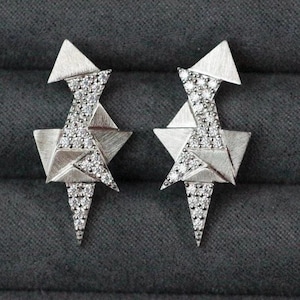 May include: A pair of silver geometric earrings. Each earring has a triangular top, a star-shaped centre, and a pointed bottom, all embellished with small, sparkling stones. The earrings have a modern, angular design.