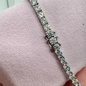 May include: A silver tennis bracelet featuring round, clear gemstones. The bracelet is fastened and lies on a pink surface. The gemstones are set closely together, creating a continuous line of sparkle.