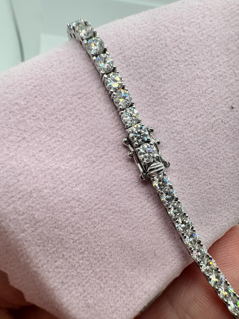 May include: A silver tennis bracelet featuring round, clear gemstones. The bracelet is fastened and lies on a pink surface. The gemstones are set closely together, creating a continuous line of sparkle.