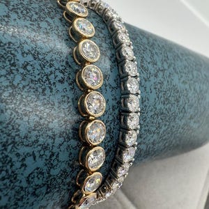 May include: Two diamond bracelets. One with oval-cut diamonds in a gold setting, the other with round diamonds in a silver setting. Both are displayed on a textured, teal surface. The bracelets are examples of fine jewellery.