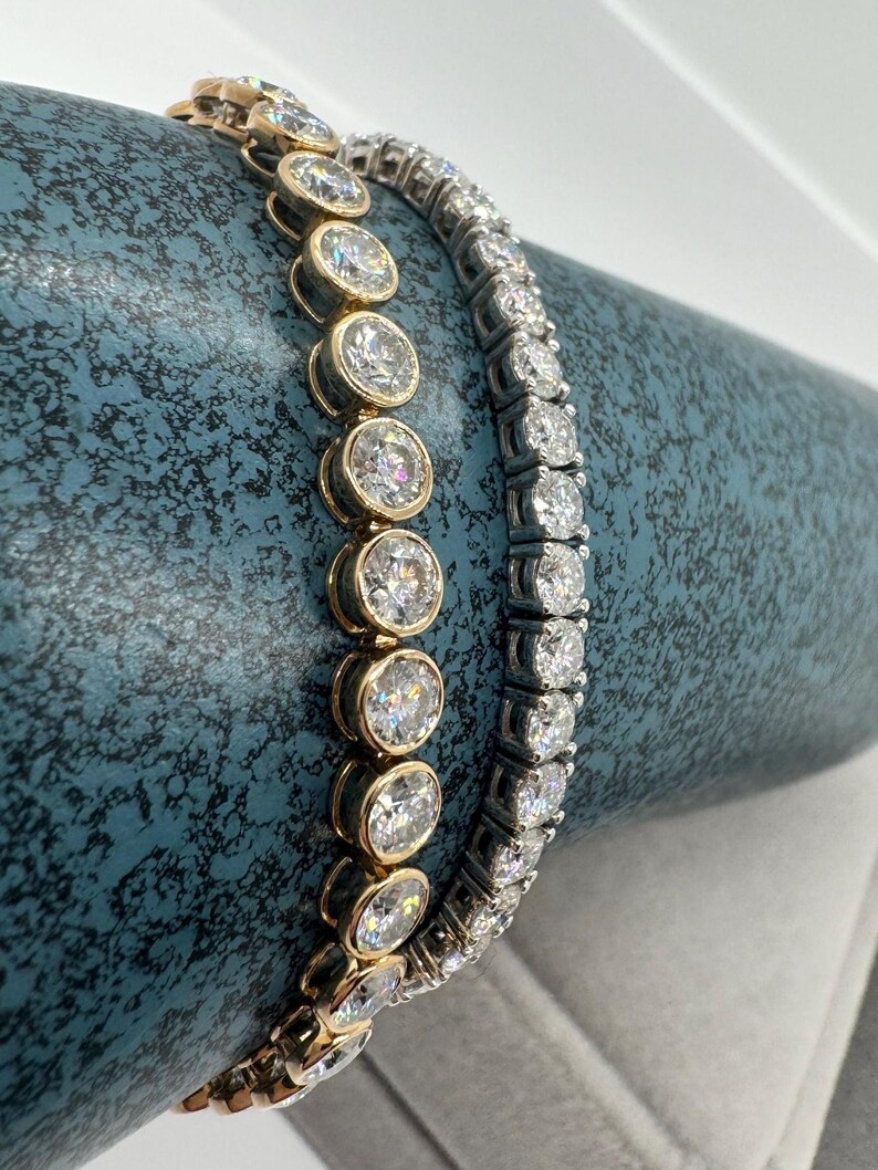 May include: Two diamond bracelets. One with oval-cut diamonds in a gold setting, the other with round diamonds in a silver setting. Both are displayed on a textured, teal surface. The bracelets are examples of fine jewellery.