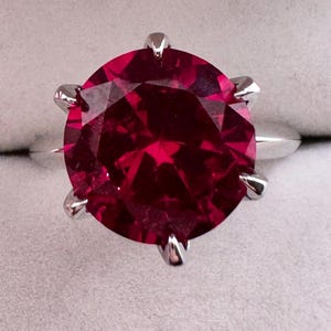 Pode incluir: Um close-up de um anel de rubi. O anel apresenta uma grande pedra preciosa redonda, facetada, de cor vermelha escura, engastada numa banda prateada. A pedra preciosa é mantida no lugar por seis garras.