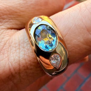May include: A gold ring featuring a large, oval-cut, light blue gemstone set in the center, flanked by two smaller, pear-shaped, clear white gemstones. The band is wide, smooth, and polished, with a rounded profile that tapers toward the back.