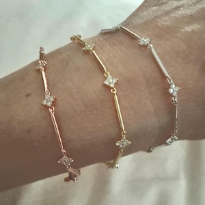 May include: Three delicate bracelets in rose gold, gold, and silver. Each bracelet features a series of small, sparkling, four-petal flower-shaped accents connected by thin bars. The bracelets are displayed on a wrist.