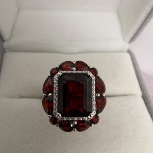 May include: A silver ring featuring a large, rectangular, dark red gemstone at its centre, encircled by smaller, oval-shaped red gemstones. The central stone is framed by a row of small, clear stones. The ring is presented in a grey jewellery box.