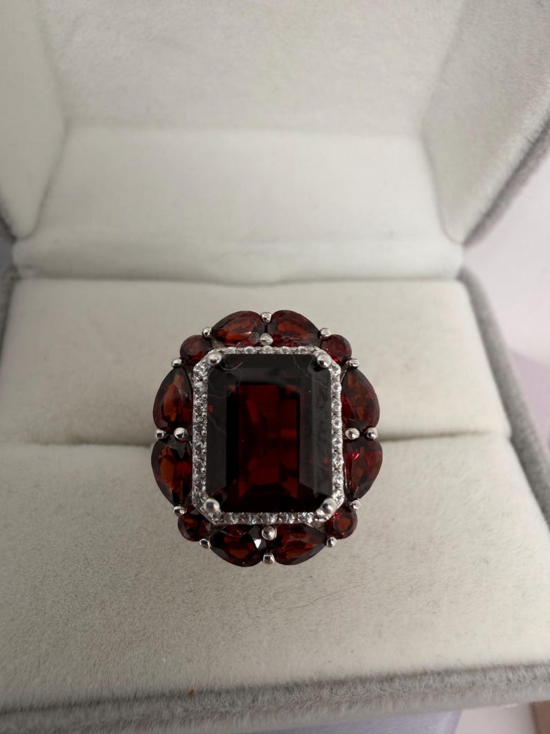 May include: A silver ring featuring a large, rectangular, dark red gemstone at its centre, encircled by smaller, oval-shaped red gemstones. The central stone is framed by a row of small, clear stones. The ring is presented in a grey jewellery box.