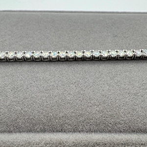 May include: A silver tennis bracelet featuring numerous round, clear gemstones set closely together. The bracelet is presented on a grey velvet surface, which contrasts with the sparkle of the stones. The bracelet is a piece of jewellery.
