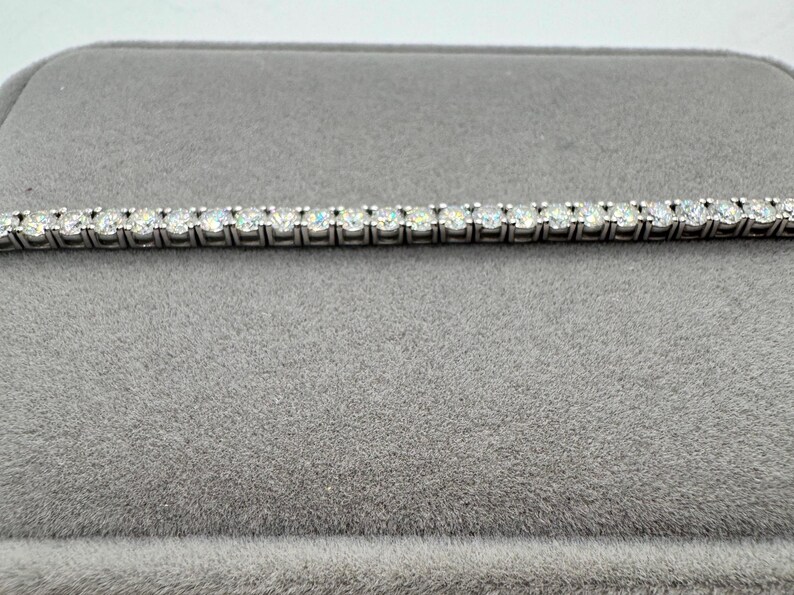 May include: A silver tennis bracelet featuring numerous round, clear gemstones set closely together. The bracelet is presented on a grey velvet surface, which contrasts with the sparkle of the stones. The bracelet is a piece of jewellery.
