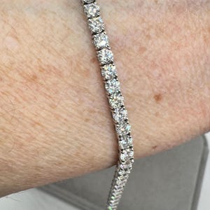 May include: A tennis bracelet with a line of round-cut diamonds set in a silver metal. The bracelet is shown on an arm, highlighting the sparkle of the gemstones. A timeless piece of jewellery.
