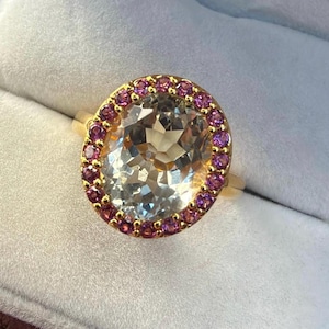 May include: A gold ring featuring a large, oval-cut, pale translucent gemstone at the center, surrounded by a halo of small, round, vibrant pink stones. The setting is crafted from polished yellow gold, highlighting the contrast between the light center stone and the bright pink border.
