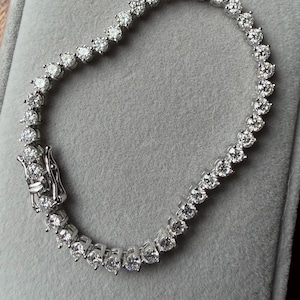 May include: A tennis bracelet featuring numerous round-cut diamonds set in a silver metal. The diamonds are closely set together, creating a continuous sparkle. The bracelet is displayed on a gray surface.