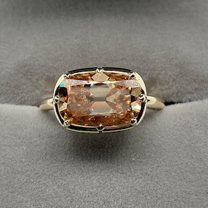 May include: A gold ring featuring a large, rectangular, light-brown gemstone. The gemstone is set in a gold bezel with small decorative accents. The ring is displayed against a gray background, highlighting the ring's design and color.