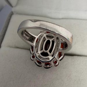 May include: A silver ring with a large, oval, dark red gemstone at its centre, encircled by smaller red stones. The ring has an intricate, openwork design around the central stone.