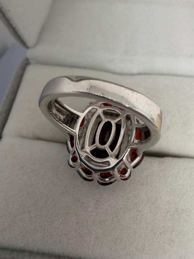 May include: A silver ring with a large, oval, dark red gemstone at its centre, encircled by smaller red stones. The ring has an intricate, openwork design around the central stone.