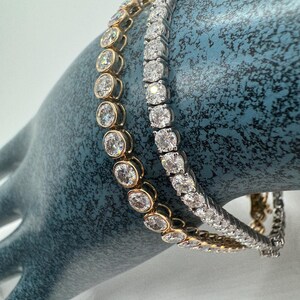 May include: Two diamond tennis bracelets. One bracelet features round, bezel-set diamonds in a gold setting. The other bracelet has square-cut diamonds in a silver setting. Both bracelets are displayed on a blue surface.