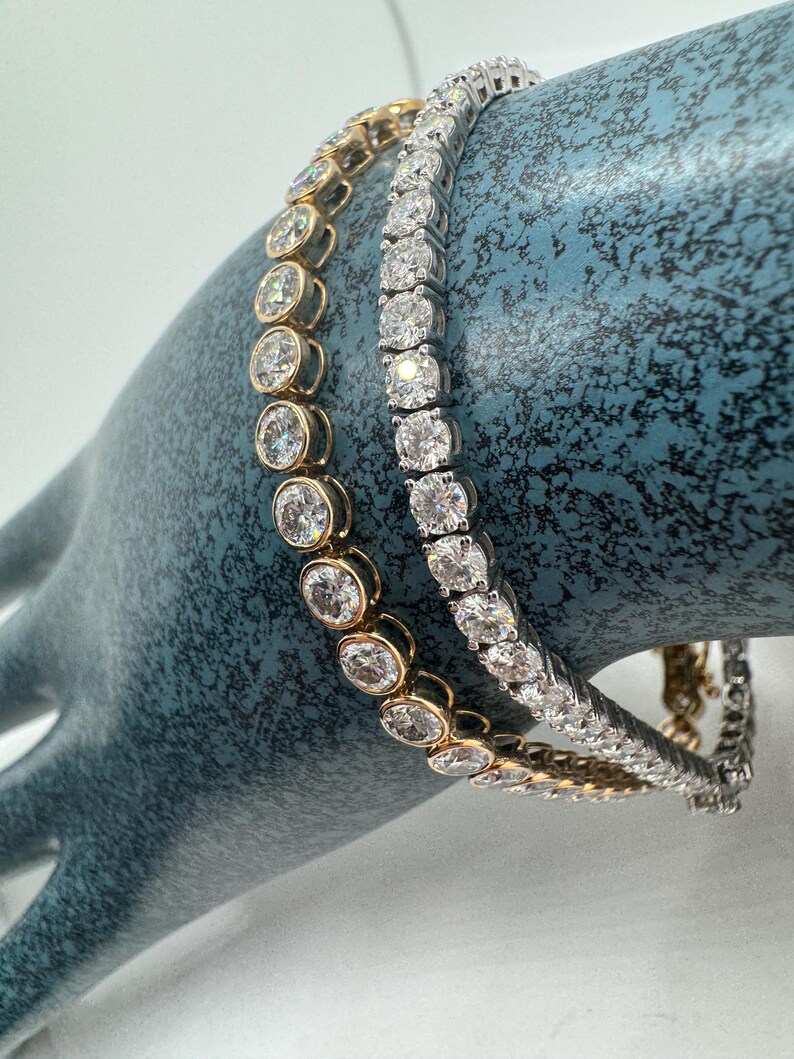 May include: Two diamond tennis bracelets. One bracelet features round, bezel-set diamonds in a gold setting. The other bracelet has square-cut diamonds in a silver setting. Both bracelets are displayed on a blue surface.