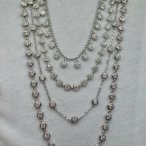 May include: A silver necklace with multiple strands of small round stones. The necklace has a layered look with a cascading effect.