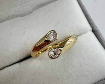 May include: A gold-colored ring with a bypass design, featuring two pear-shaped clear gemstones. The ring is displayed inside a gray jewelry box, showcasing its polished finish and elegant style.