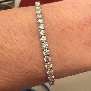 May include: A tennis bracelet with a row of round-cut diamonds set in a silver or white gold band. The diamonds are sparkling. The bracelet is displayed on a person's arm.