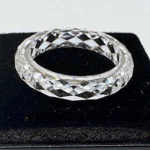 May include: A clear, faceted ring with a geometric design. The ring is made of a transparent material, likely crystal or glass, and sits on a black surface. The ring's surface is cut with many small, angled facets.
