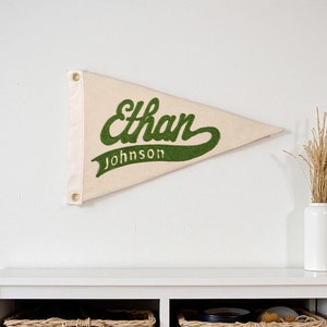 May include: A cream-coloured pennant displaying the name "Ethan Johnson" in green script. The triangular pennant has reinforced eyelets for hanging. The design is simple and classic, suitable for a bedroom or office.