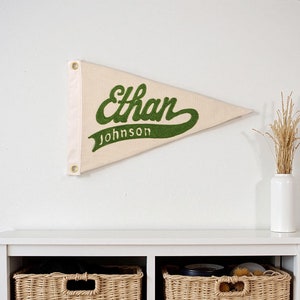 Könnte beinhalten: Ein weißer Wimpel mit grüner Beschriftung, auf dem "Ethan Johnson" steht. Der Wimpel hängt an einer weißen Wand über einem weißen Regal mit zwei Weidenkörben.