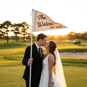 May include: A couple kissing on a golf course at sunset. The groom wears a black suit and the bride a white wedding dress. A white flag with the text "The Waters EST. 2026" is held above them.