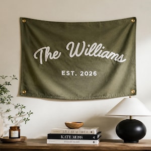 Könnte beinhalten: Ein olivgrünes Wandbanner aus Stoff mit dem weißen gestickten Text The Williams und EST. 2026. Das rechteckige Banner hat vier messingfarbene Metallösen an den Ecken zum Aufhängen und eine strukturierte, gewebte Oberfläche.
