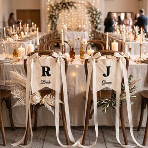 Op de afbeelding: Bruiloftdecoratie met twee houten stoelen met witte banners met de tekst "Bride" en "Groom". De banners zijn versierd met witte linten. De tafel is gedekt met kaarsen, bloemen en gouden accenten, wat een romantische sfeer creëert.