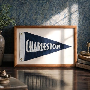 Op de afbeelding: Een driehoekige marineblauwe viltwimpel met de witte geborduurde tekst Charleston, gemonteerd in een rechthoekige houten lijst. De wimpel heeft een witte verticale strook aan de linkerkant met twee messingkleurige metalen zeilringen.