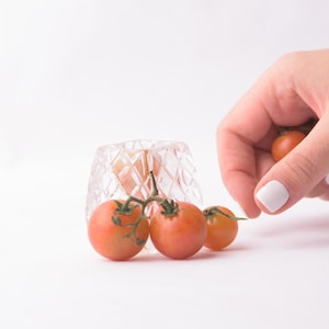 Könnte beinhalten: Ein klarer Glasbehälter mit Diamantmuster und einer Traube reifer, roter Cherrytomaten an einer Rebe. Eine Hand mit weißem Nagellack greift nach einer Tomate. Die Tomaten befinden sich auf weißem Hintergrund.